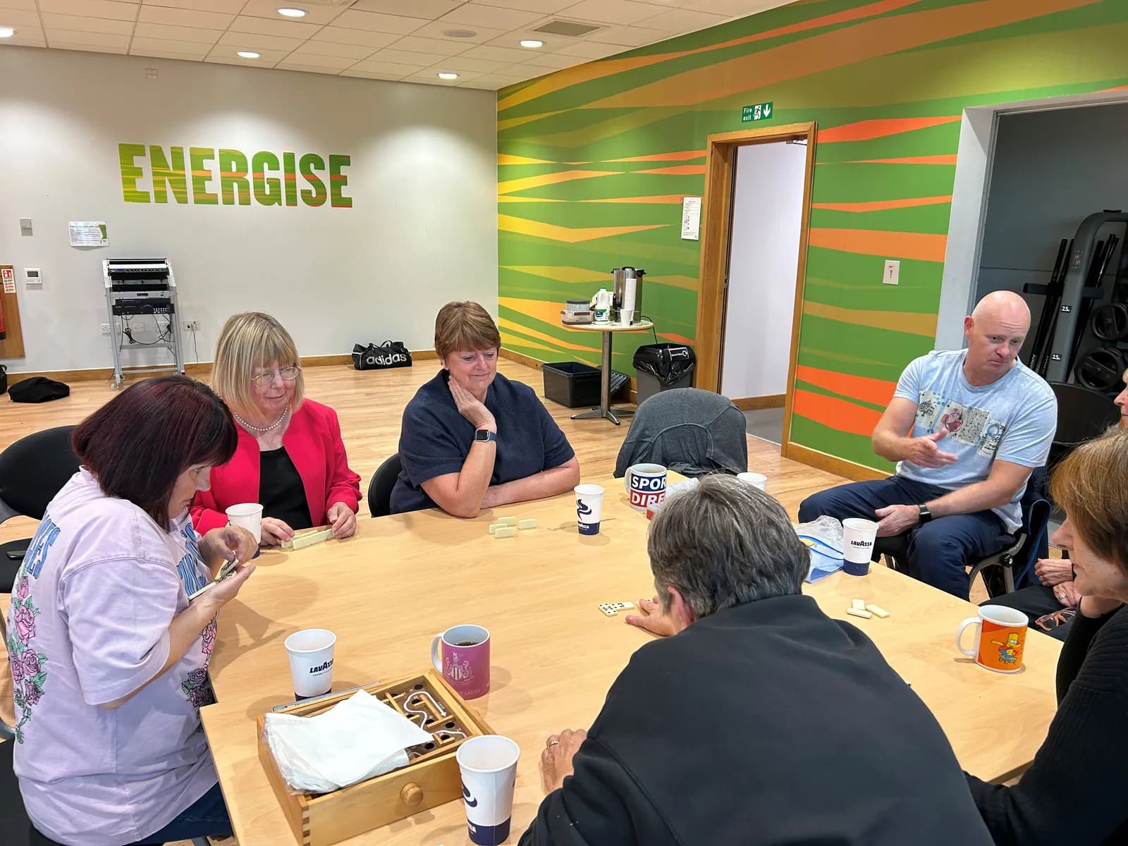 A group of adults sits around a light wooden table playing a game, with a large green and orange wall behind them that reads "ENERGISE".
